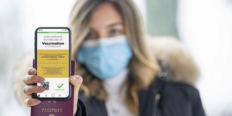 A defocused young girl, wearing a face mask, is holding a passport and a smart phone with a with an example of a certificate of vaccination against the covid 19 disease.