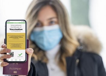 A defocused young girl, wearing a face mask, is holding a passport and a smart phone with a with an example of a certificate of vaccination against the covid 19 disease.